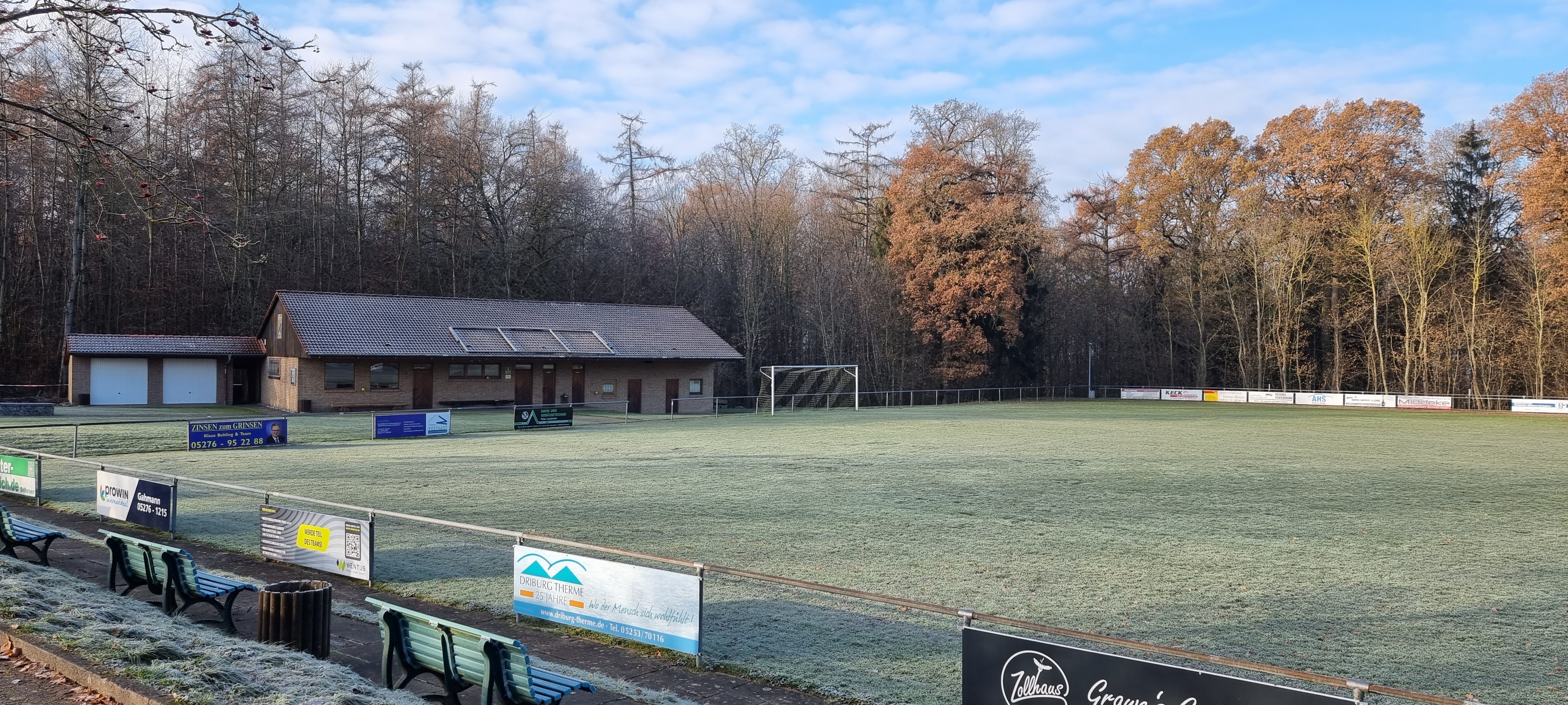 Das Bild zeigt einen Sportplatz mit Tribüne, Absperrzaun und Gebäude.