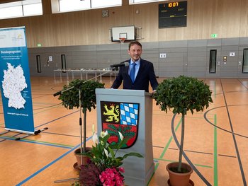 Stadt Weiden Das Bild zeigt einen Mann im Anzug hinter einem Rednerpult mit Wappen in einer Turnhalle.
