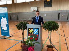 Das Bild zeigt einen Mann im Anzug hinter einem Rednerpult mit Wappen in einer Turnhalle.