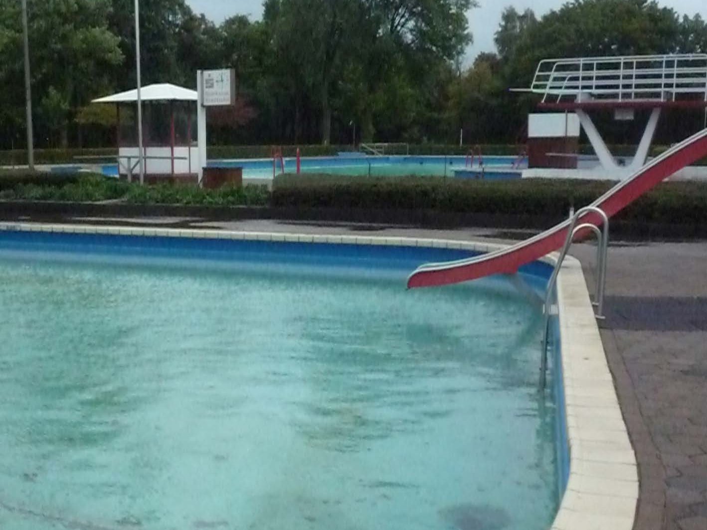 Das Bild zeigt ein leeres Freibadbecken mit Rutsche und Sprungturm vor der Sanierun