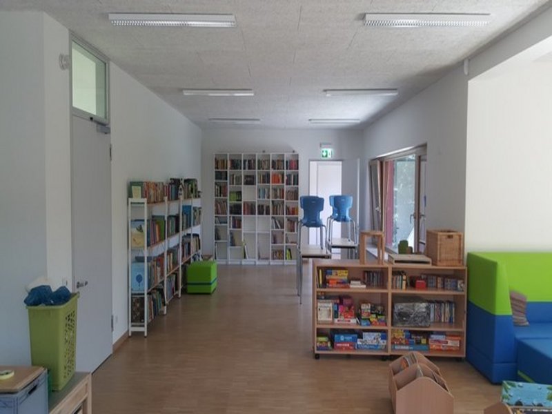 Das Bild zeigt einen hellen Raum mit Regalen voller Bücher und Spiele sowie grünen und blauen Möbelelementen.