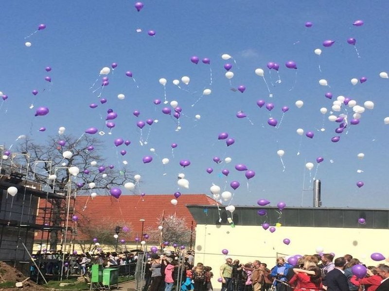 Das Bild zeigt lila und weiße Luftballons, die über Menschen am Boden hinweg in den Himmel steigen.