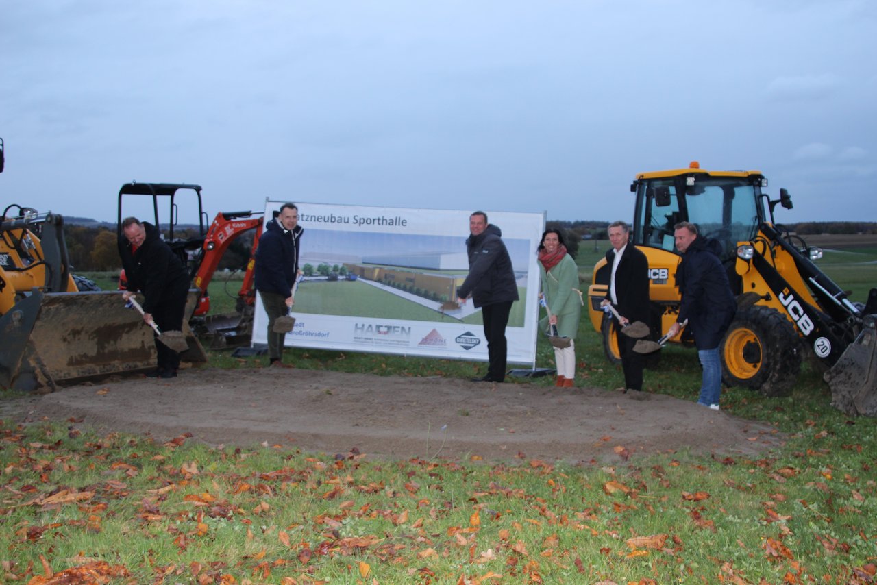 Das Bild zeigt 6 Personen mit Spaten vor Baumaschinen auf freier Wiese.