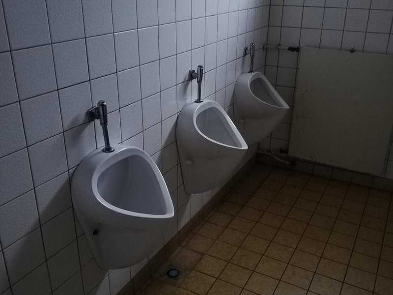 Man sieht drei Urinale an einer gefliesten Wand.
