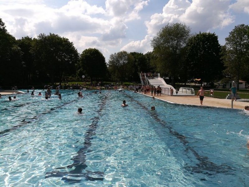 Das Bild zeigt ein Schwimmbecken mit einigen Menschen, im Hintergrund ist eine Wellenrutsche zu sehen.