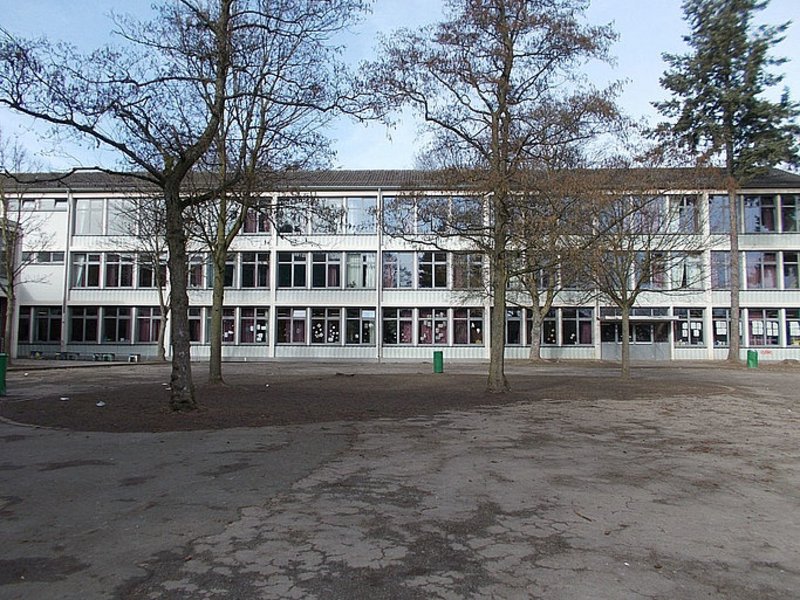 Das Bild zeigt ein fensterreiches Gebäude, im Vordergrund ist ein Platz mit Bäumen.