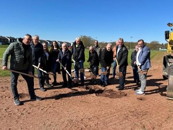 Das Bild zeigt 13 Personen, die nebeneinander in einer Reihe stehen und alle einen Spaten halten. 