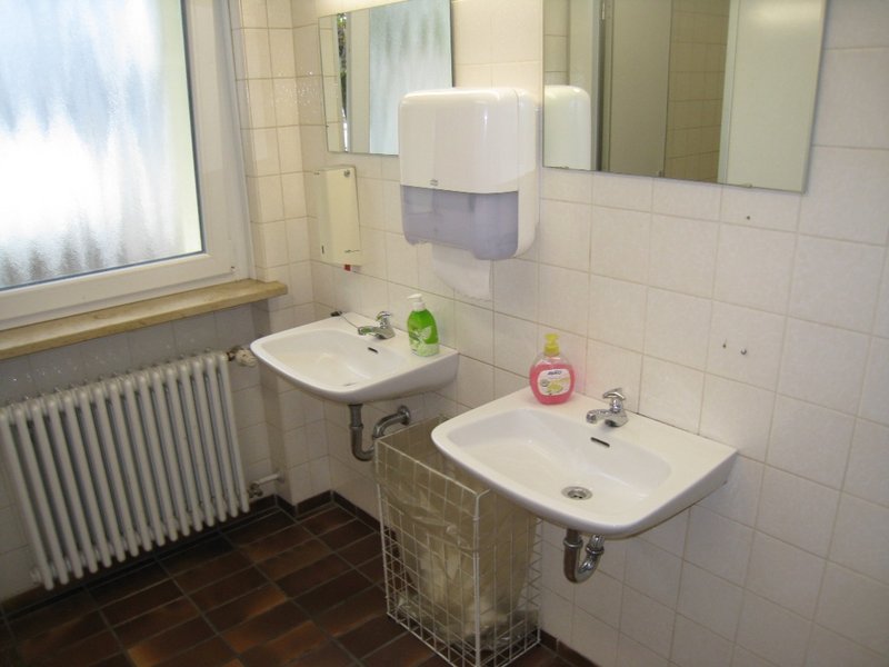 Man sieht den Waschbereich einer Toilette mit gefliesten Wänden und zwei Spiegeln über zwei Waschbecken.