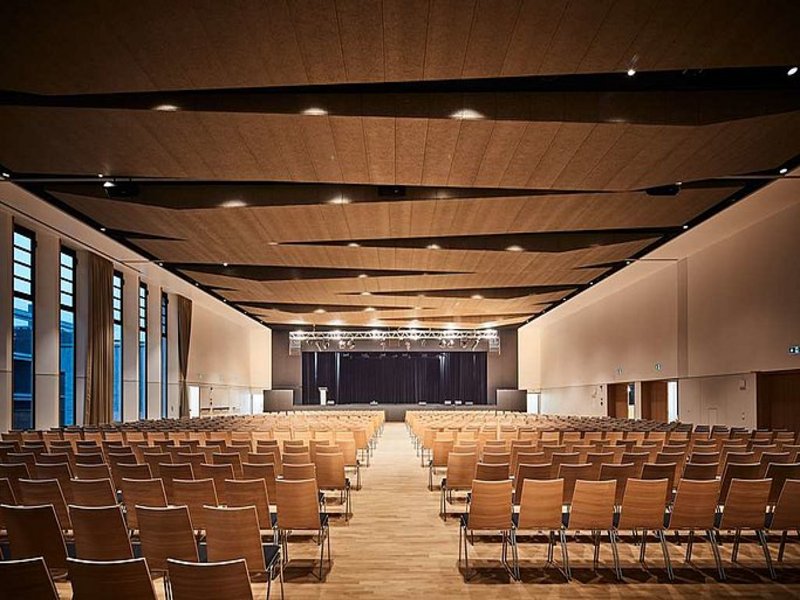 Das Bild zeigt einen großen, hell erleuchteten Saal mit Stuhlreihen und einer Bühne.
