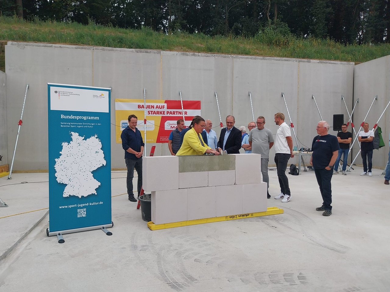 Das Bild zeigt ein Stück Mauer, um das mehrere Personen versammelt sind. Eine Frau legt eine Zeitkapsel in die Mauer.