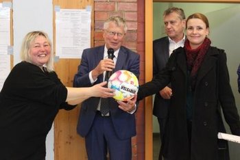 Das Bild zeigt zwei Frauen und einen Mann mit Mikrofon, die die Hände auf einen Fußball gelegt haben. Im Hintergrund steht ein weiterer Mann.