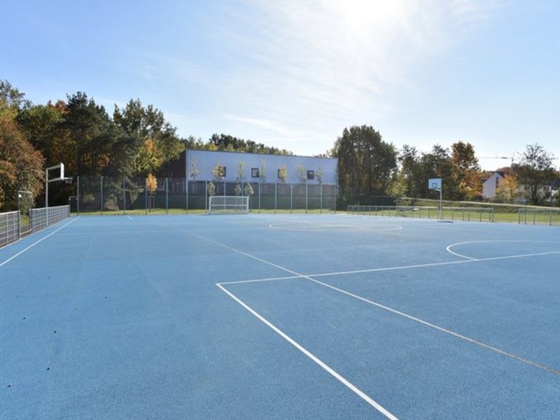 Das Bild zeigt eine Sportaußenanlage mit blauem Boden und weißen Markierungen, im Hintergrund ist ein weißes Gebäude mit Flachdach zu sehen.