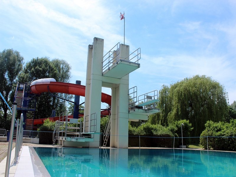 Das Bild zeigt den Sprungturm und eine rote Rutsche vor einem Schwimmbecken.