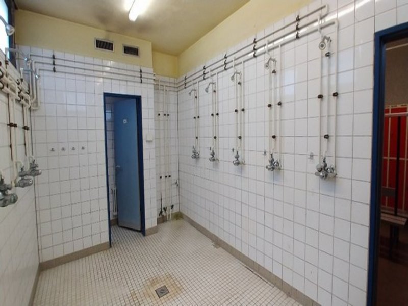 Das Bild zeigt einen in die Jahre gekommenen Sanitärbereich mit weißen Fliesen und mehreren Duschen.