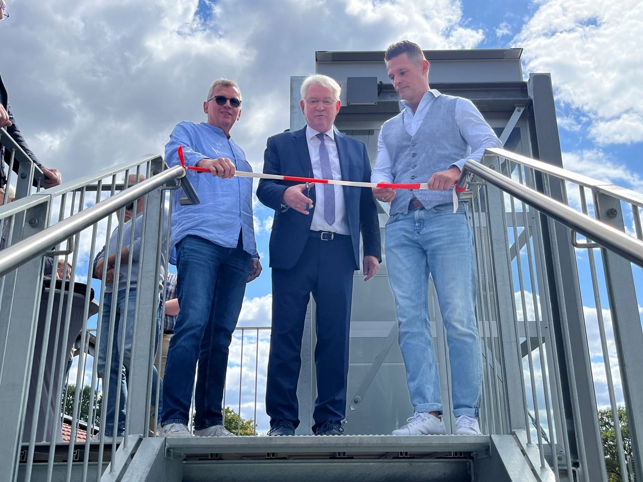 Quelle: Gemeinde Augustdorf Das Bild zeigt drei Männer auf der oberen Schwelle einer Außentreppe, einer von ihnen schneidet ein Band durch.