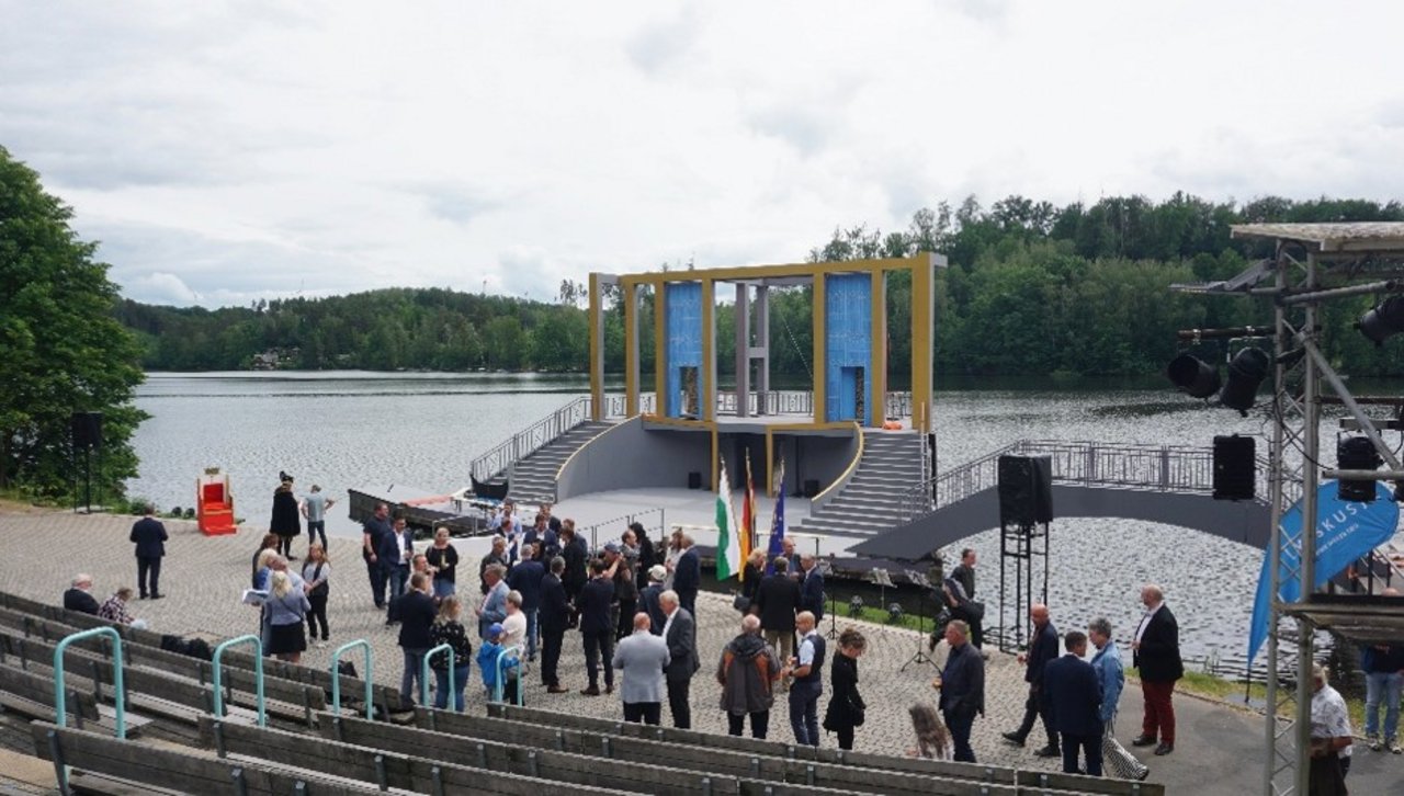 Quelle: Keine Angabe Das Bild zeigt die feierliche Einweihung der neuen Seebühne in Kriebstein. Man erkennt eine Menschenmenge um Lausprecher verteilt. Im Hintergrund befidet sich eine Bühne auf dem See.