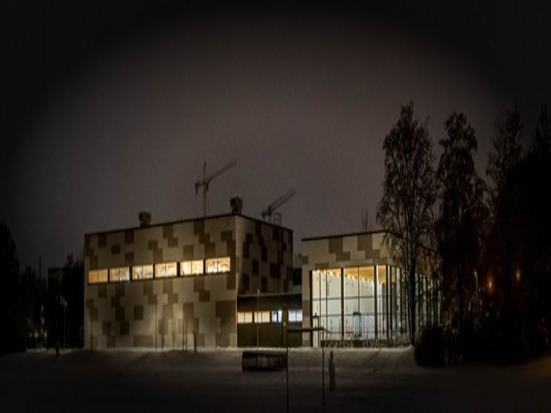 Das Bild zeigt ein beleuchtetes Gebäude mit Flachdach, Fensterfronten und bunter Fassade bei Nacht.