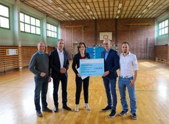Das Bild zeigt eine Gruppe von Menschen in einer Sporthalle. Zwei von ihnen halten gemeinsam einen großen Förderscheck in Händen.