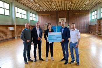 Das Bild zeigt eine Gruppe von Menschen in einer Sporthalle. Zwei von ihnen halten gemeinsam einen großen Förderscheck in Händen. 