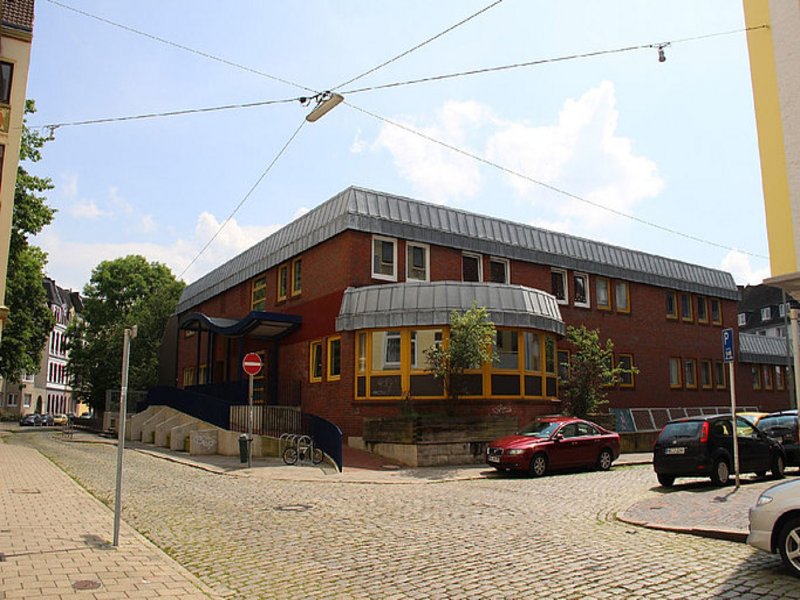 Das Bild zeigt ein Gebäude aus Backsteinen mit gelben Fenstern und einem halbrunden Anbau im Erdgeschoss.