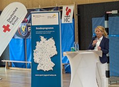 Das Bild zeigt eine Frau mit Mikrofon an einem Stehtisch, im Hintergrund sind mehrere Aufsteller und Sportgeräte zu sehen.