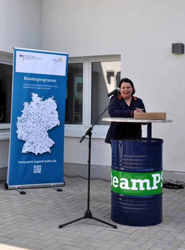 Das Bild zeigt eine Frau, die vor einem einer Hauswand an einem Stehtisch aus einer Tonne in ein Mikrofon spricht. 