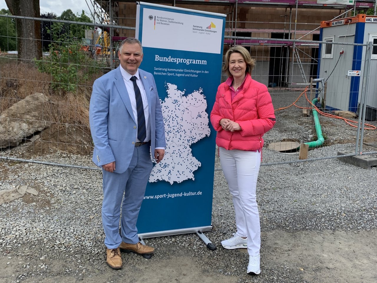 Das Bild zeigt 2 Personen, in der Mitte von ihnen ist ein Roll-Up zu sehen, im Hintergrund befindet sich eine Baustellensituation.