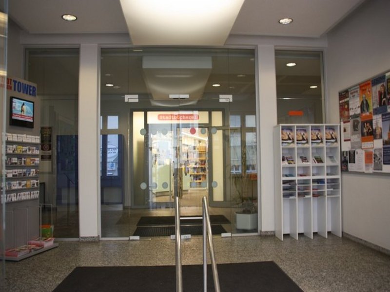 Das Bild zeigt den Eingang zu einer Stadtbücherei mit Glastüren und Prospektständern.