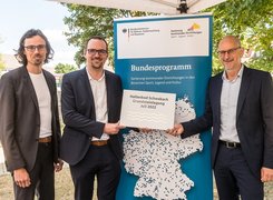 Man sieht drei Männer vor dem Roll Up zum Bundesprogramm bei der Grundsteinlegung.