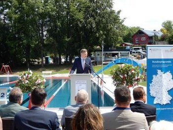 Das Bild zeigt einen Mann, der vor einem Publikum eine Rede hält. Im Hintergrund ist das Schwimmerbecken eines Freibades zu sehen.