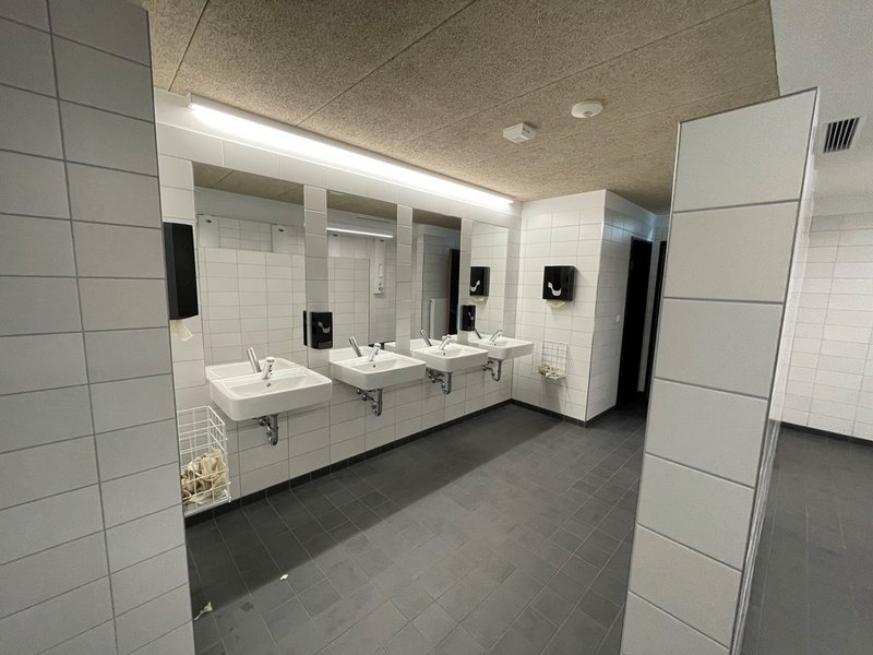 Das Bild zeigt vier Waschbecken in einer Reihe mit einer Spiegelfront, der Boden ist grau gefliest, die Wände weiß.