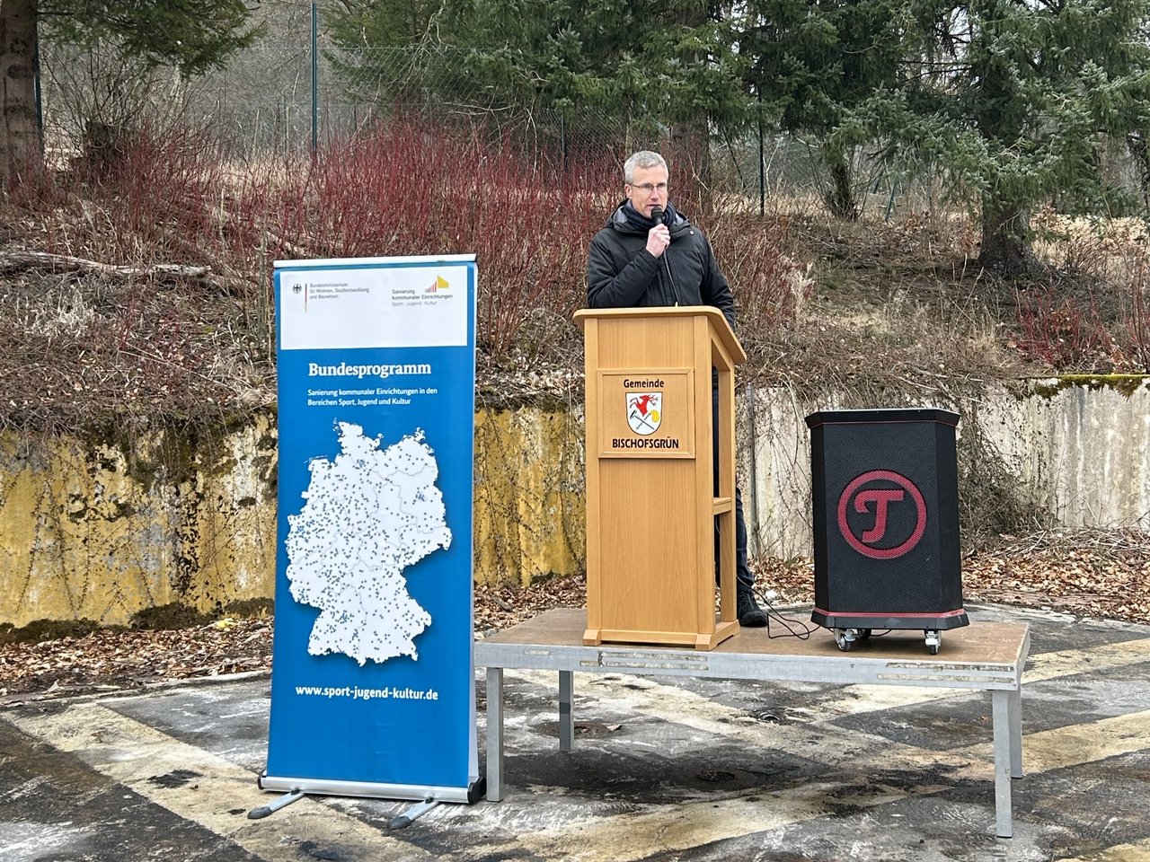 Das Bild zeigt ein Redepult mit Redner bei dem Spatenstich. Im Vordergrund steht ein Roll-Up.
