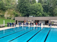 Das Bild zeigt im Vordergrund ein Schwimmbecken, im Hintergrund stehen Menschen vor einem Gebäude versammelt.