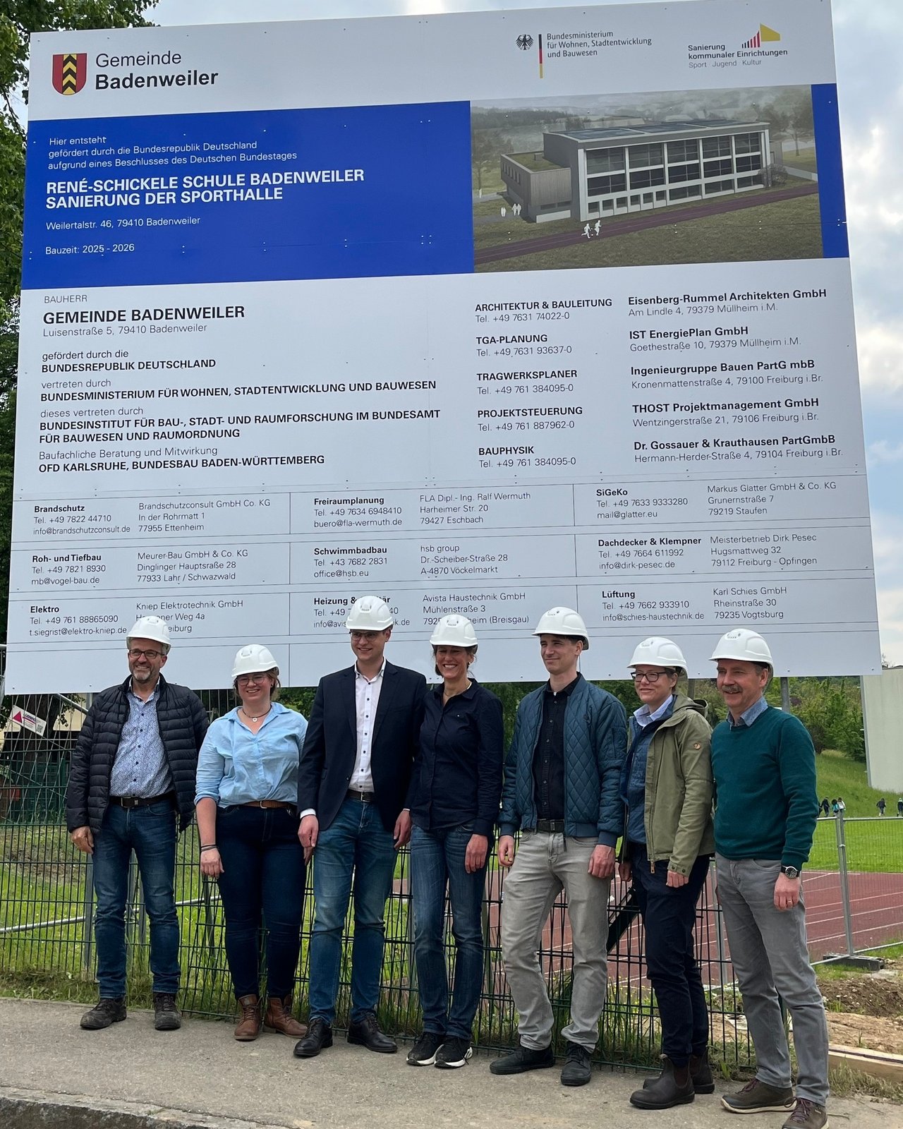 Auf dem Bild sind eine Gruppe Menschen vor einer Tafel zum Projekt zu sehen, die symbolisch einen Baustellenhelm tragen.