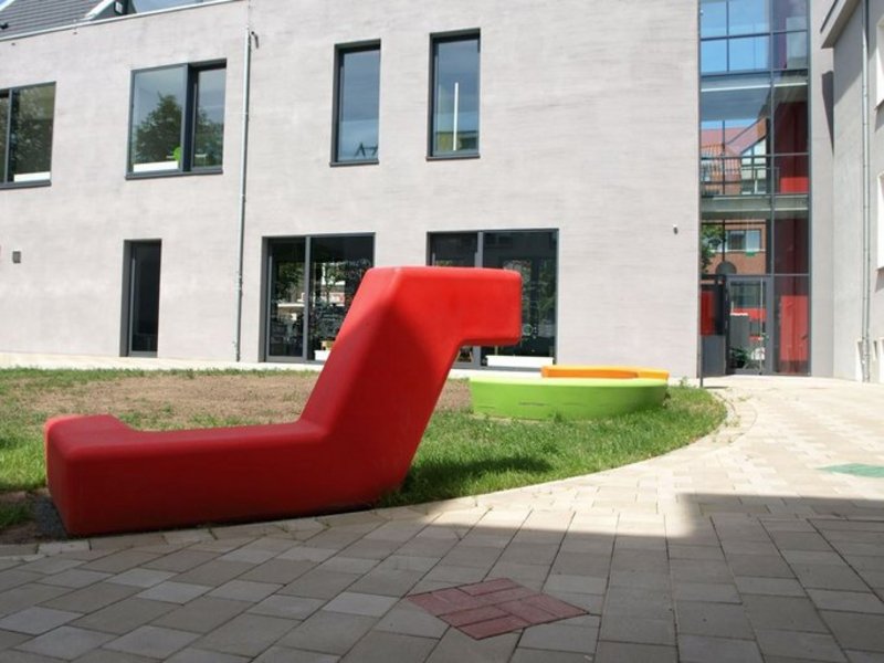 Das Bild zeigt eine rote Sitzgelegenheit am Übergang von Pflaster auf Rasen.
