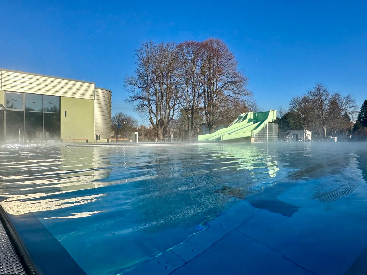 Quelle: Bäder, Sport und Freizeit Salzgitter GmbH Das Bild zeigt ein Außenschwimmbecken mit einer Rutsche und einem Gebäude im Hintergrund.