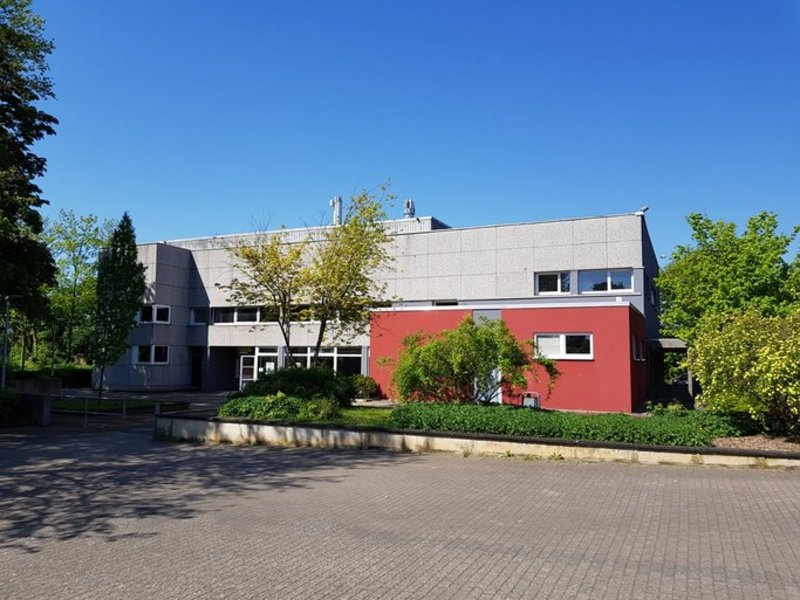 Das Bild zeigt ein Flachdach-Gebäude mit roter Fassade im Erdgeschoss und grauer Fassade im 1. Obergeschoss, umgeben von Bäumen.