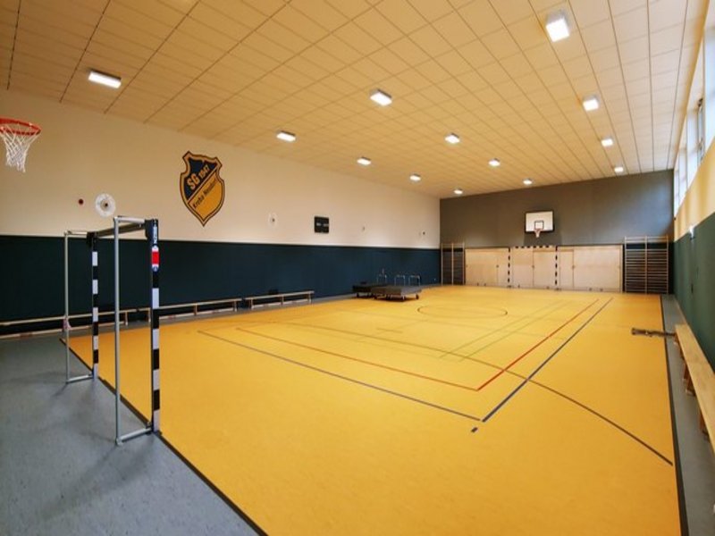 Das Bild zeigt eine Sporthalle mit gelbem Boden, Fußballtor, Basketballkorb und Vereinslogo an der Wand.