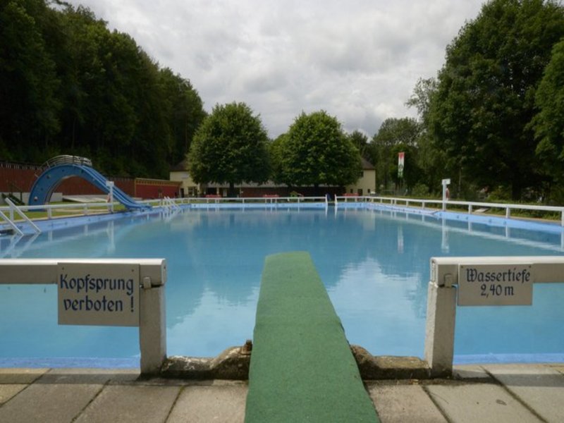 Das Bild zeigt im Vordergrund Startblöcke, im Hintergrund ist ein Schwimmbecken im Außenbereich erkennbar.