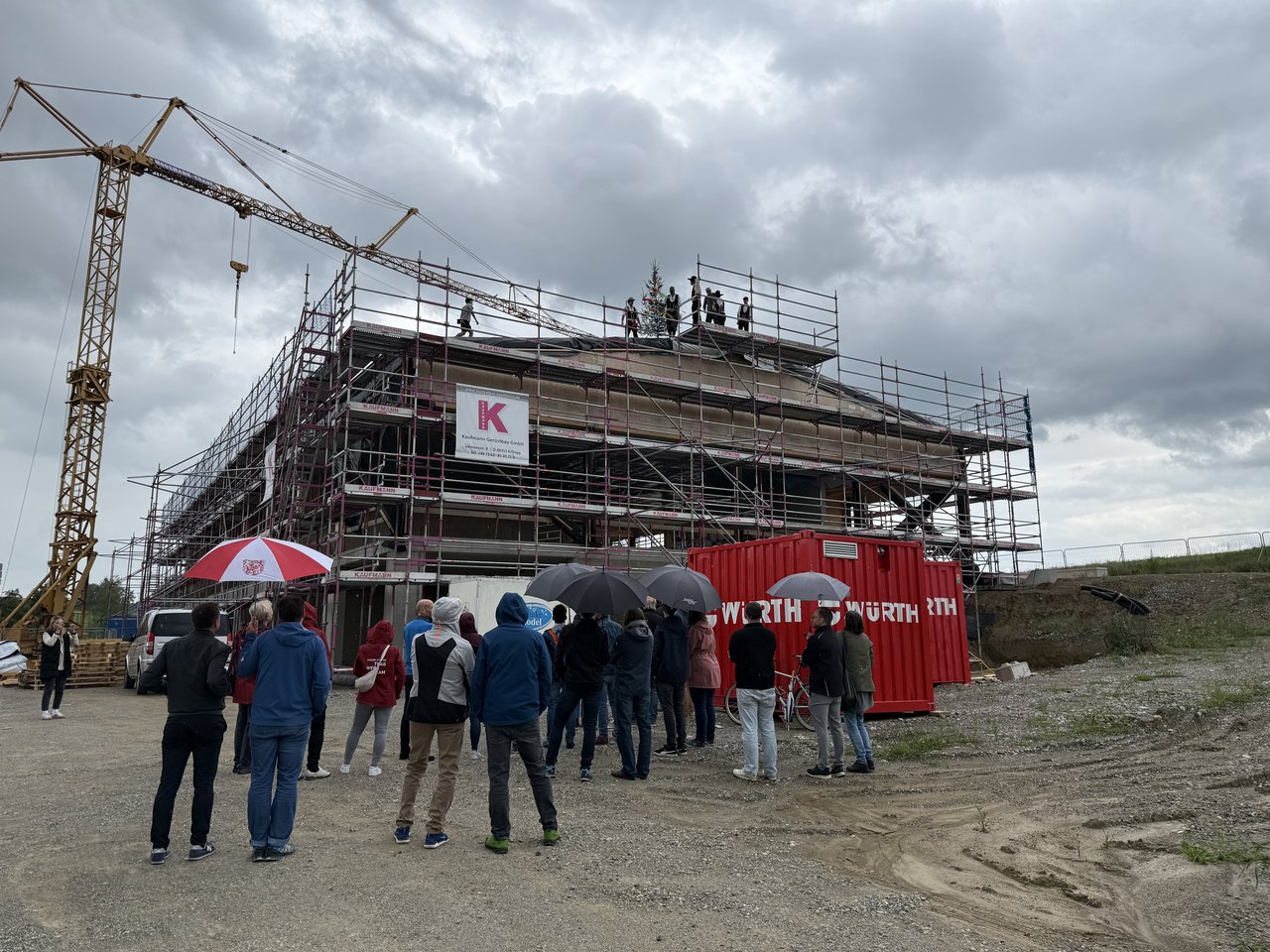 Das Bild zeigt eine Baustelle mit einem Rohbau, vor dem mehrere Personen mit Regenschirmen stehen. Auf dem Dach stehen ebenfalls einige Personen.