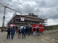 Das Bild zeigt eine Baustelle mit einem Rohbau, vor dem mehrere Personen mit Regenschirmen stehen. Auf dem Dach stehen ebenfalls einige Personen.