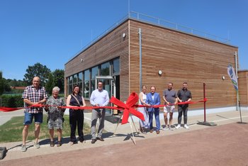 Acht Personen stehen vor einem Gebäude mit Holzfassade und durchtrennen gemeinsam ein rotes Band mit mehreren Scheren.