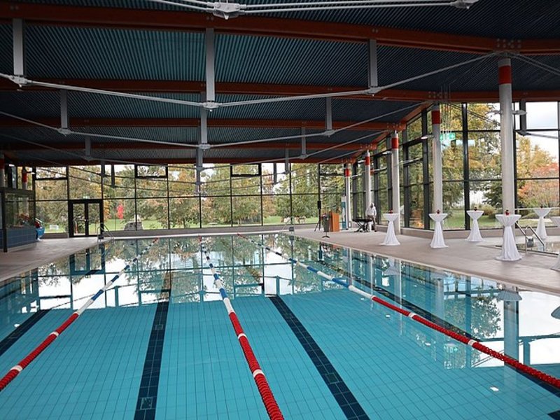 Das Bild zeigt ein menschenleeres Schwimmbecken in einem Hallenbad, am Beckenrand sind Stehtische mit weißen Hussen zu sehen.