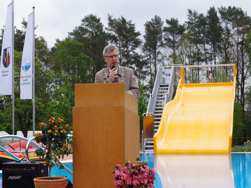 Das Bild zeigt einen Mann an einem Rednerpult in einem Freibad, im Hintergrund sind ein Becken und eine gelbe Wellenrutsche zu sehen.