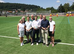 Das Bild zeigt acht Personen bei der Einweihung einer Sportanlage. Im Hintergrund wird Fußball gespielt.