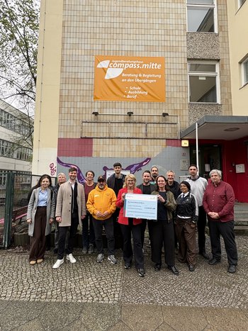 Eine Gruppe Menschen steht vor einem Gebäude und posiert für ein Foto. Zwei Personen in der Mitte halten einen Förderscheck in der Hand.