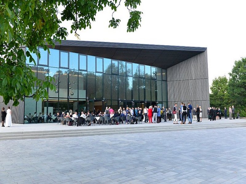Das Bild zeigt ein modernes Gebäude, auf dessen Vorplatz Menschen stehen und sitzen.