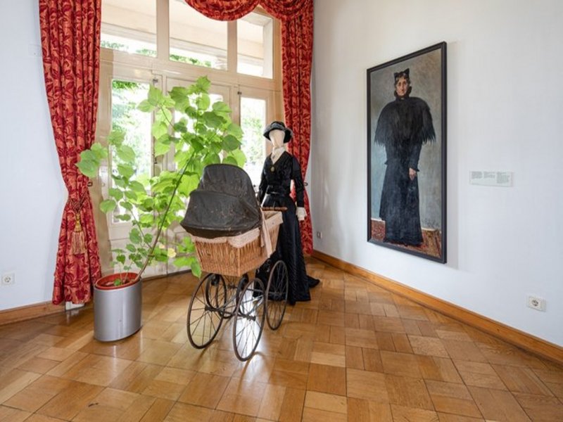 Das Bild zeigt einen Innenraum mit Parkett, es ist eine Puppe in historischem Gewand mit Kinderwagen, ein Gemälde an der Wand, eine Pflanze sowie im Hintergrund ein Fenster mit schweren roten Vorhängen zu sehen.