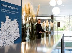 Das Bild zeigt eine Frau hinter einem Rednerpult. Daneben ist ein Roll-Up zu sehen, auf dem eine Deutschlandkarte abgebildet ist und Bundesprogramm Sanierung kommunaler Einrichtungen in den Bereichen Sport, Jugend und Kultur zu lesen ist, sowie Blumendekoration.