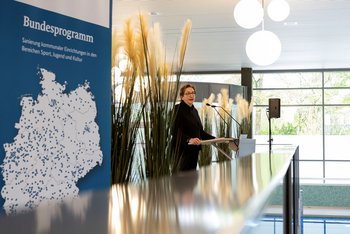 © Christina Czybik / bundesfoto Das Bild zeigt eine Frau hinter einem Rednerpult. Daneben ist ein Roll-Up zu sehen, auf dem eine Deutschlandkarte abgebildet ist und Bundesprogramm Sanierung kommunaler Einrichtungen in den Bereichen Sport, Jugend und Kultur zu lesen ist, sowie Blumendekoration.